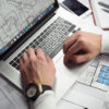 Hände arbeiten am Laptop mit technischen Bauplänen und Architekturzeichnungen auf dem Schreibtisch