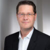 Björn Gödde in dunklem Anzug und weißem Hemd mit Brille vor neutralem Hintergrund